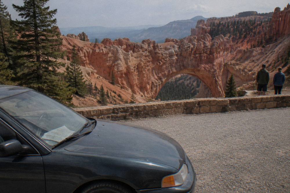 A viewpoint pullout along a rim road with pine forest and layered red rock amphitheater below