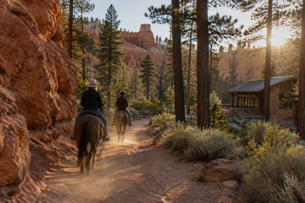 Two riders on horseback crossing a sandy forest trail flanked by red rock walls at golden hour