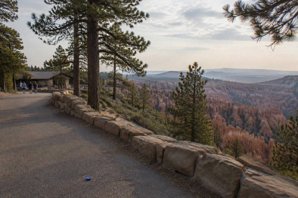 A paved overlook path along the rim with pine trees and distant eroded rock spires