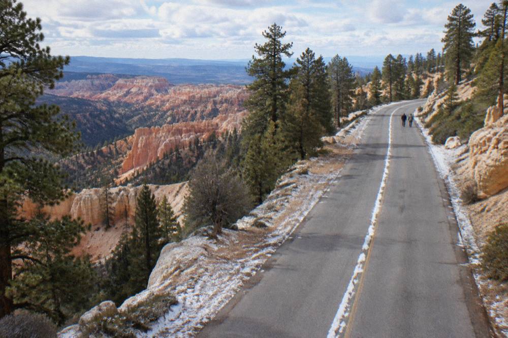 A high desert plateau road winding between pale rock formations and green ponderosa pines under scattered clouds