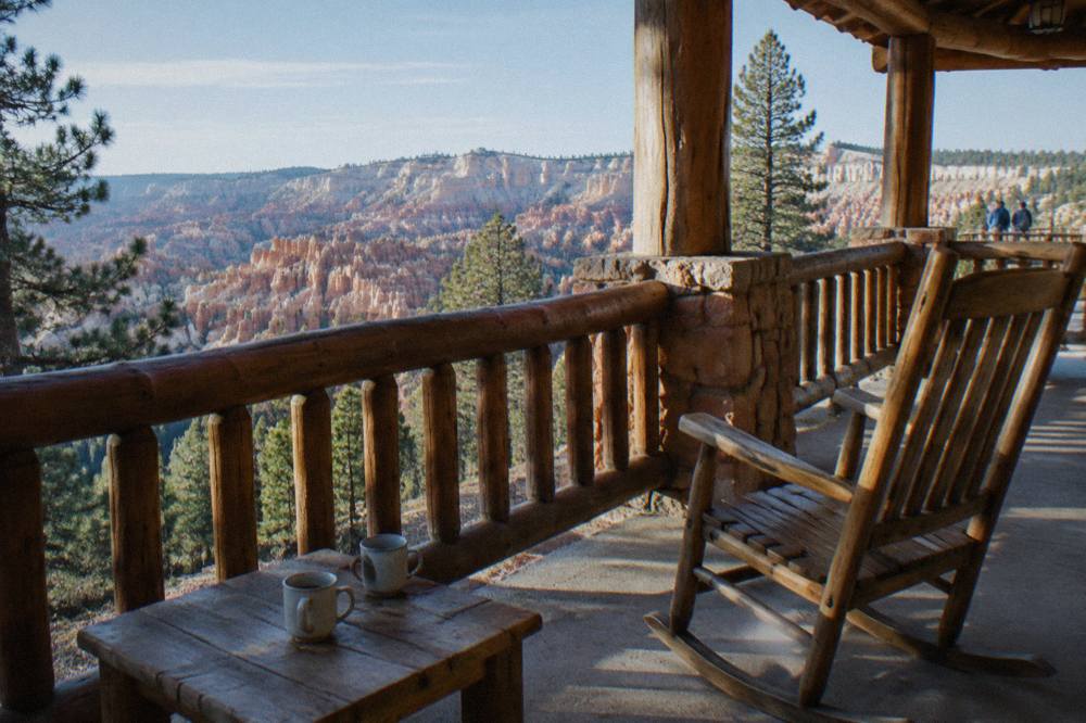 Wooden lodge veranda overlooking a pine forest toward pale rock amphitheater formations in morning light