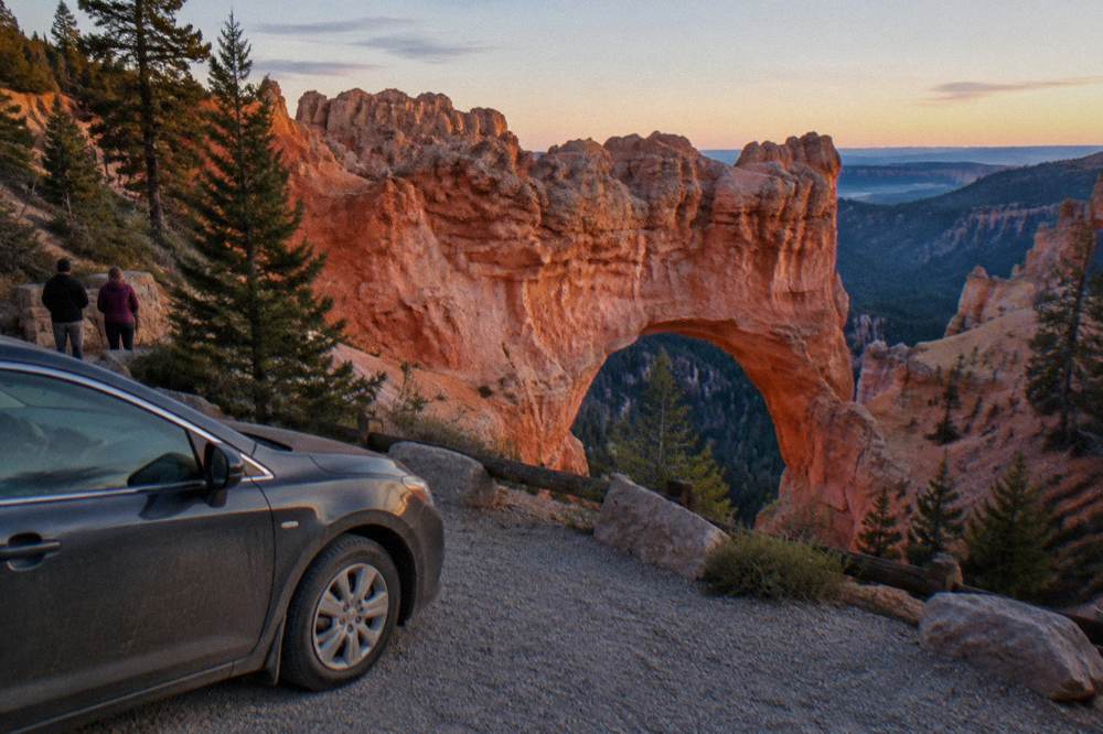 Sedan parked at a roadside overlook above layered orange rock formations at dusk