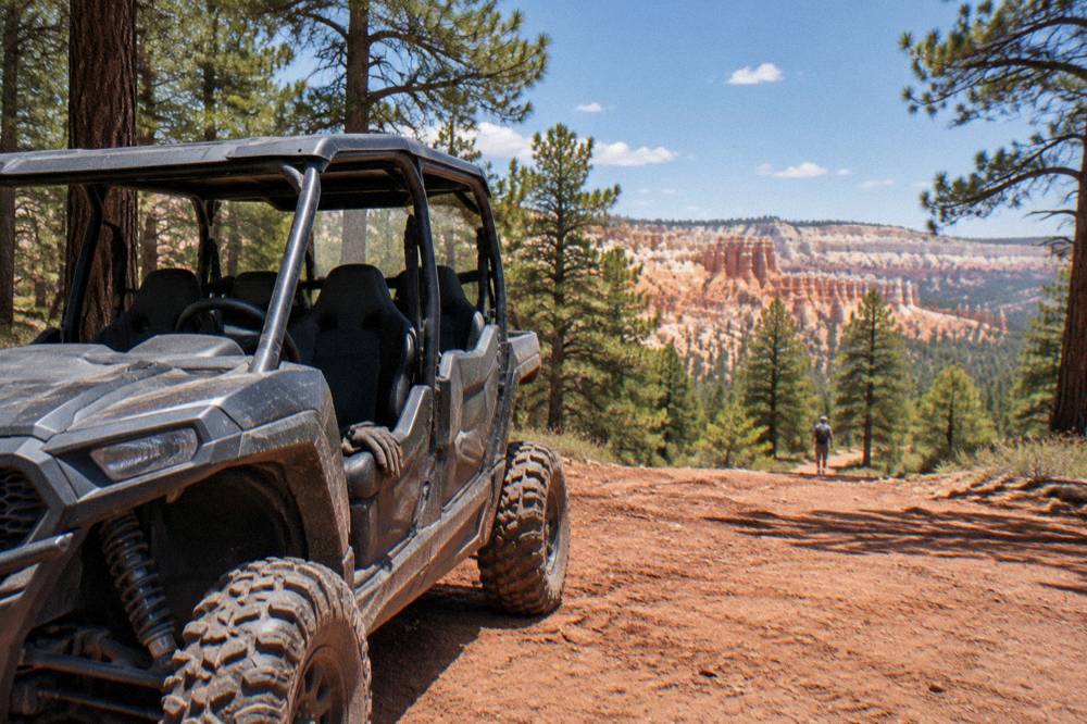 A side-by-side UTV parked on a red dirt trail with pine forest and rock cliffs behind it