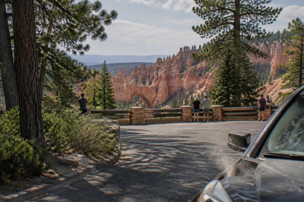 Paved overlook parking area with pine trees and a wide view of eroded red rock cliffs