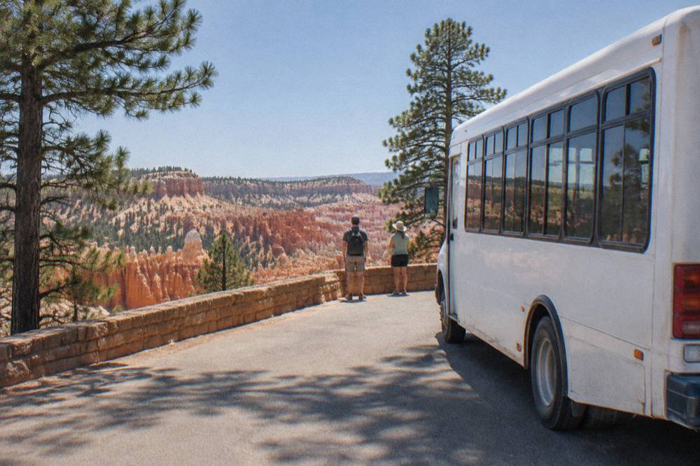 A shuttle bus at a rim overlook stop with pine forest and hoodoo formations beyond.