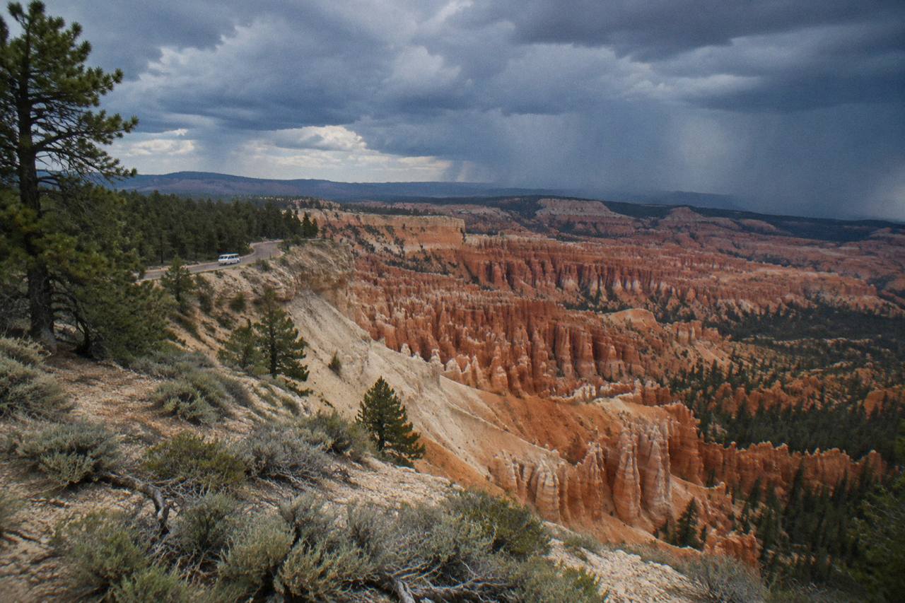 Wide from the upper overlook at Inspiration Point, Bryce Canyon National Park, Utah, August monsoon clouds stacking over the hoodoo basin, a white tour van small on the rim road below, dust snagging on sage and slickrock