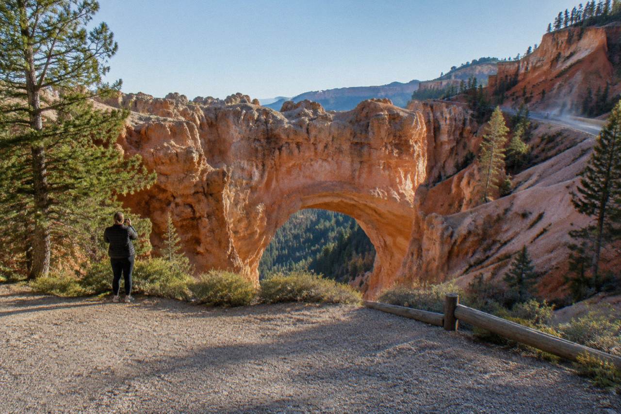 Wide view of the Natural Bridge red-rock arch on the Bryce Canyon scenic drive, Utah, pullout gravel in the foreground, afternoon sun spotlighting the arch's oxidized orange span, distant car dust drifting on the road