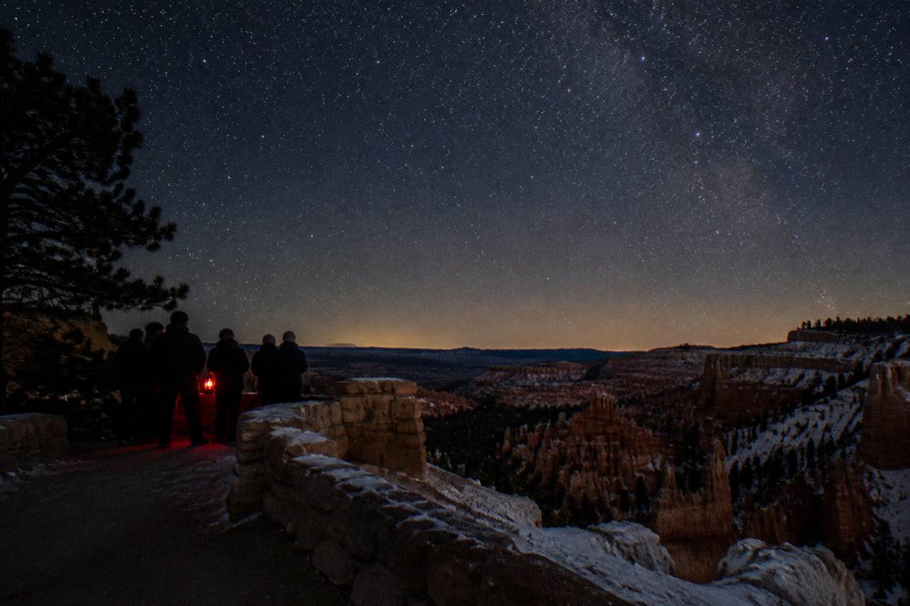 Wide night scene at Sunset Point, Bryce Canyon National Park, Utah, open sky dense with stars above the black rim, a small knot of stargazers from behind near faint red lantern light, frost glinting on the limestone rim